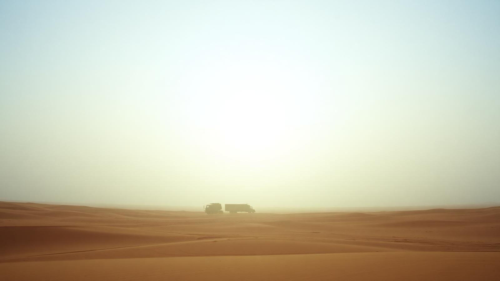 Sun-bleached desert horizon with a small convoy of military trucks under a pale sky.